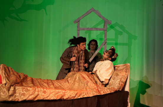 Noah Ryan as The Baker looking at Stephen Haronitis who is playing The Wolf and is in a bed, on stage in a production of "Into the Woods" at War Memorial Hall at the University of Guelph. Also on stage is a member of the ensemble observing the scene. Photo credit: Joshua Lonuzzo