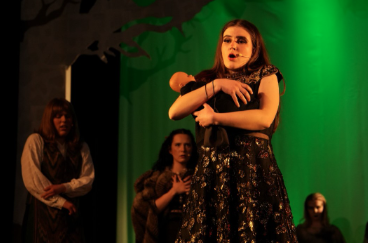  Aoibhinn Macken-Luyt as The Witch on stage in a production of "Into the Woods" at War Memorial Hall at the University of Guelph. She is holding a doll. Also on stage are Emma Harper as Jack, Charly Graham as Little Red Riding Hood and members of the ensemble. The stage is set as a forest with trees. 