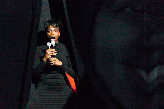 Image of Aisha Sasha John on stage in front of a black curtain with a microphone.