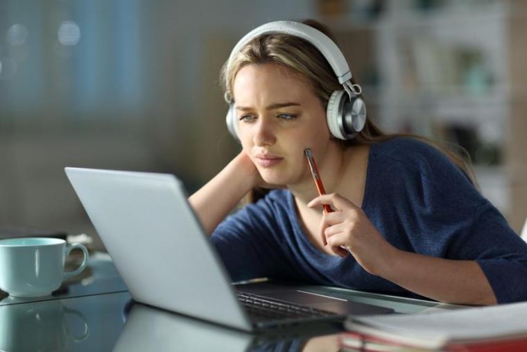 Pensive student wearing wireless headphones completing an assignment on a laptop at home.