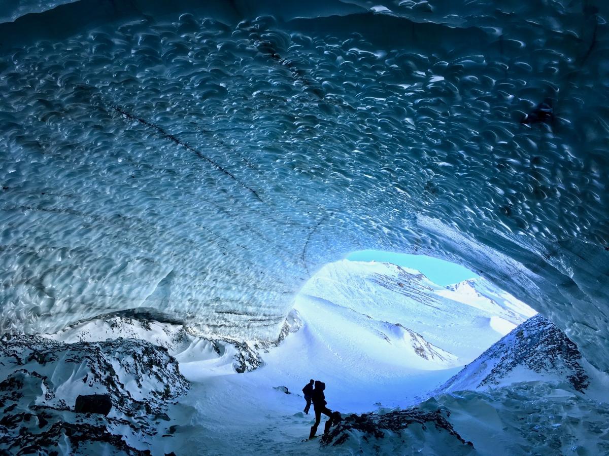 Researchers in blue ice-lined cave with snow-covered mountains in the distance.