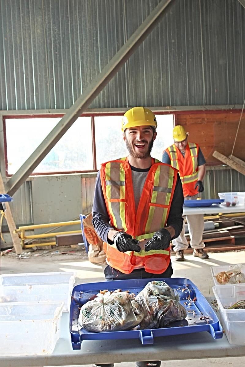 A young man smiles while sorting garbage in a warehouse with a hard hat and hi-visibility vest on.