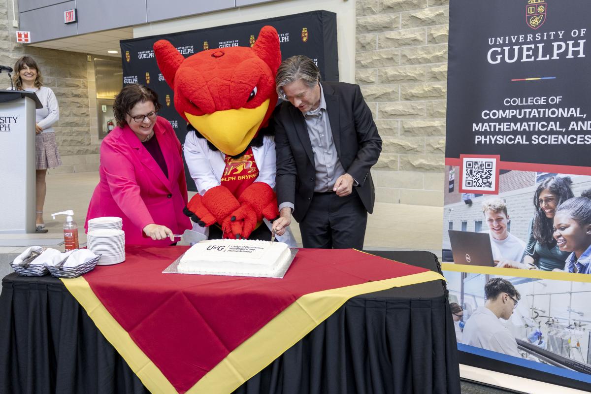 Dr. Scott, interim dean, Gryph and Dr. Rosehart cut a celebratory cake beside a CCMPS roll-up banner. Dr. Cojocaru stands to the left.