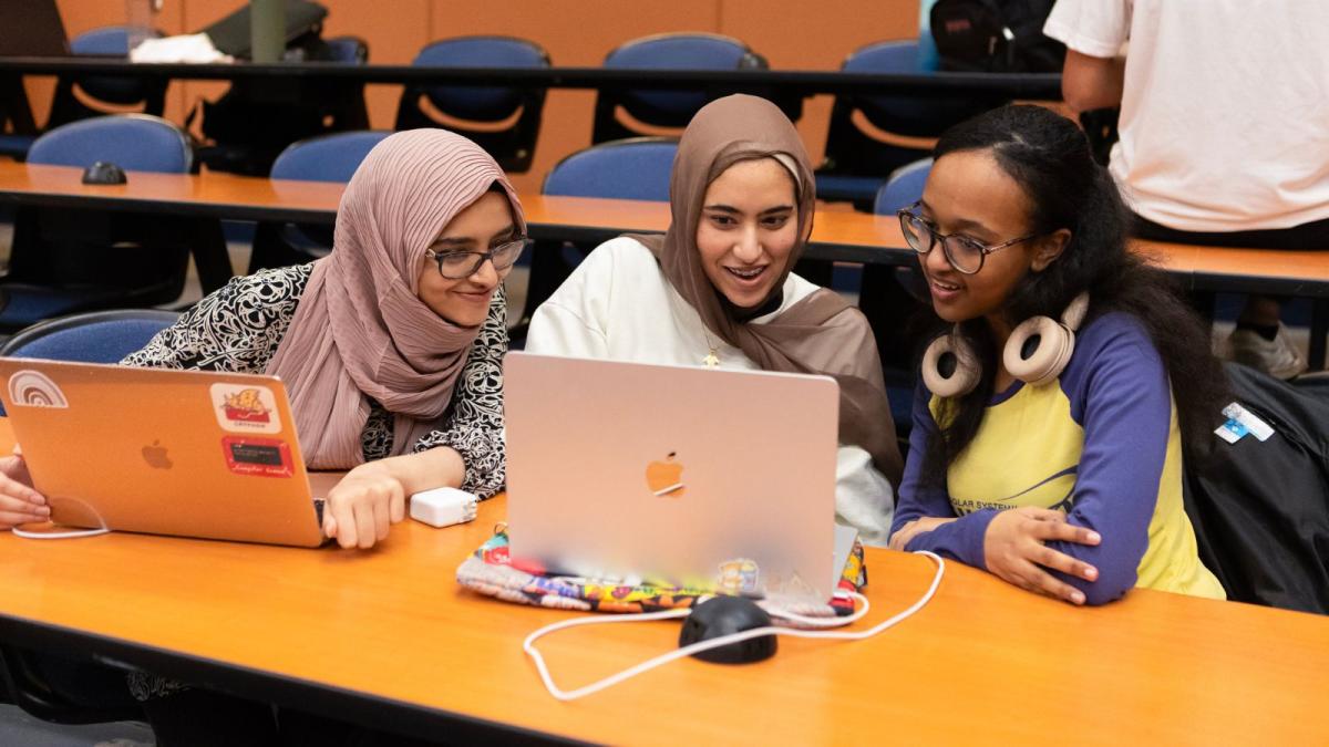 Three students around a laptop conversing. 