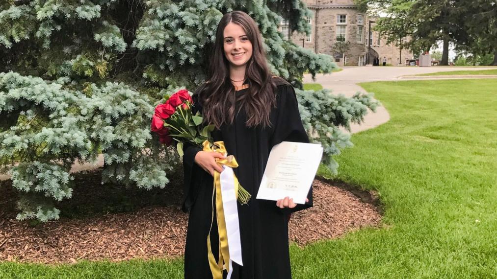 Victoria Flood smiling in her graduation gown, holding flowers and her diploma.