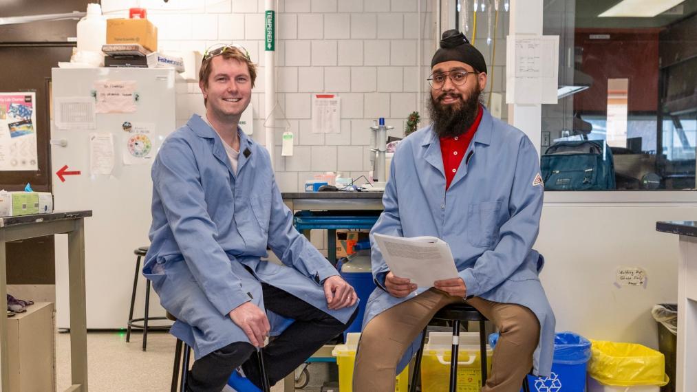  Dr. Derek K. O’Flaherty (left) sits on a stool, smiling while Jagandeep S. Saraya (right) sits on a stool, holding papers and looking toward the camera,
