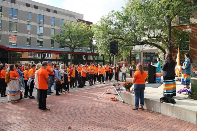 A group of people standing together wearing orange shirts listening to a speaker.