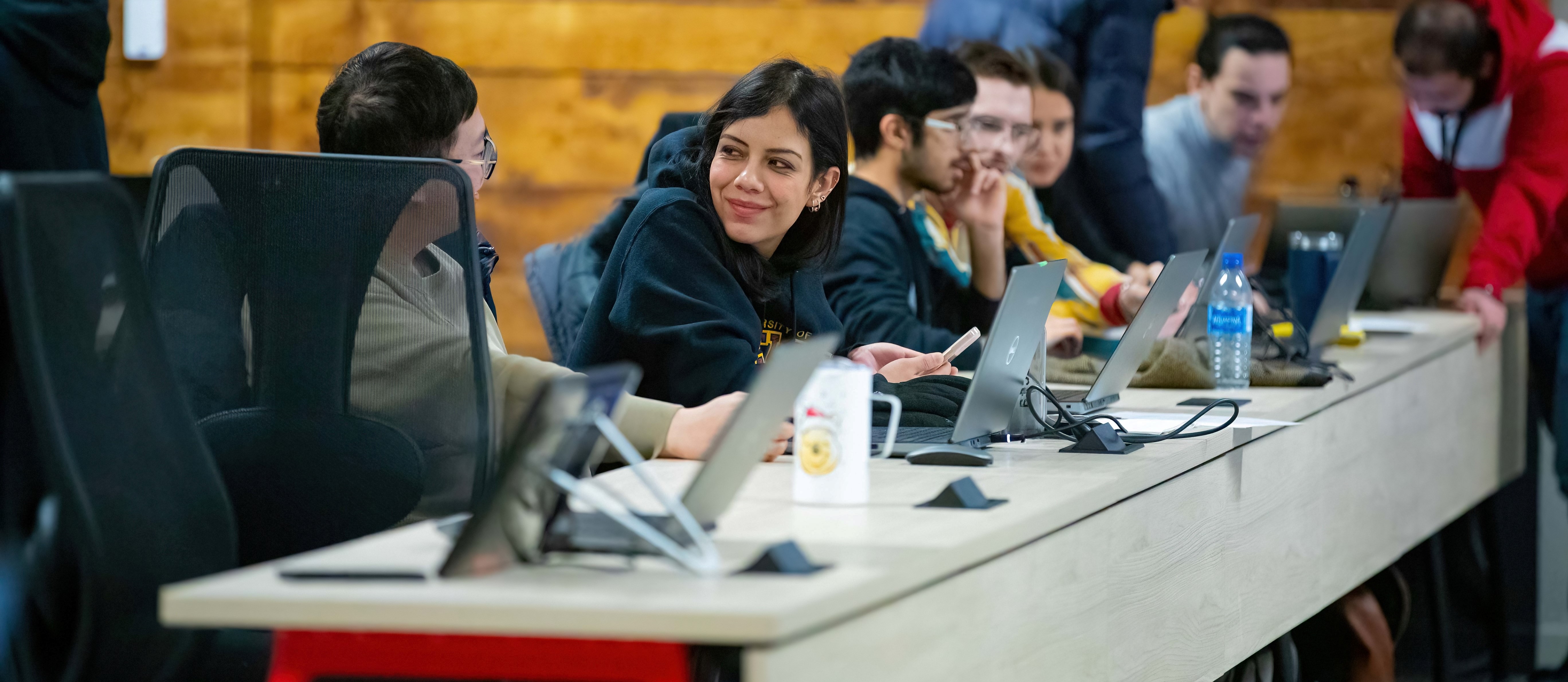 Several students sit at a long desk with their laptops as they talk to each other