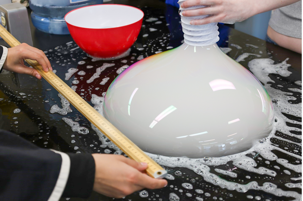 One student is holding a tube which is making a large, soapy bubble while another student is using a meter stick to measure the size of the bubble.