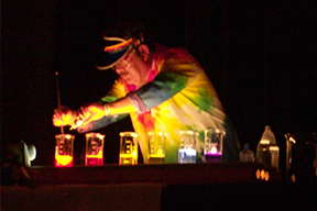 someone in a tie-dyed lab coat in dim lights in front of several beakers each filled with different coloured and glowing solutions 