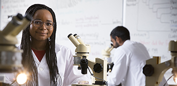 Woman wearing lab coat and looking at camera with microscope next to her