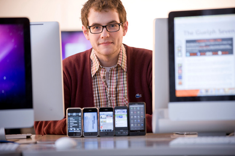 Image of student looking at camera with several mobile devices showing apps. A computer screen reads "The Guelph 7"