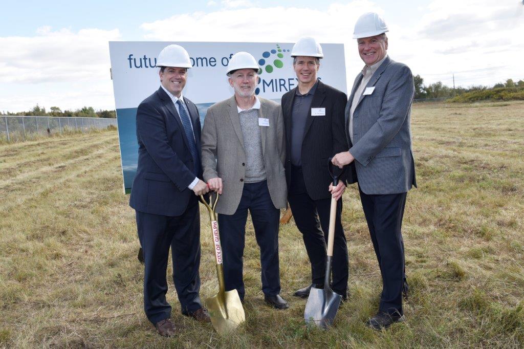 Image of four people standing in field in front of sign holding shovels