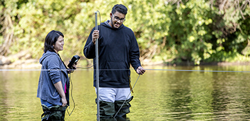 Two students wearing waders and standing in stream while looking at research equipment