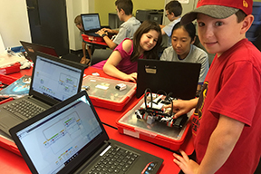 boy with electronics smiling at the camera and girls in the background working on a computer program