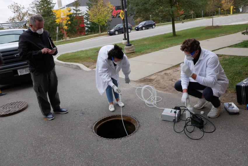 Engineering prof. Ed McBean stands to the left of two student research assistants Jonathan Evans and Melissa Novacefski. McBean is looking at data on his phone while the students are squatted over a manhole with the cover removed, using equipment to collect samples.
