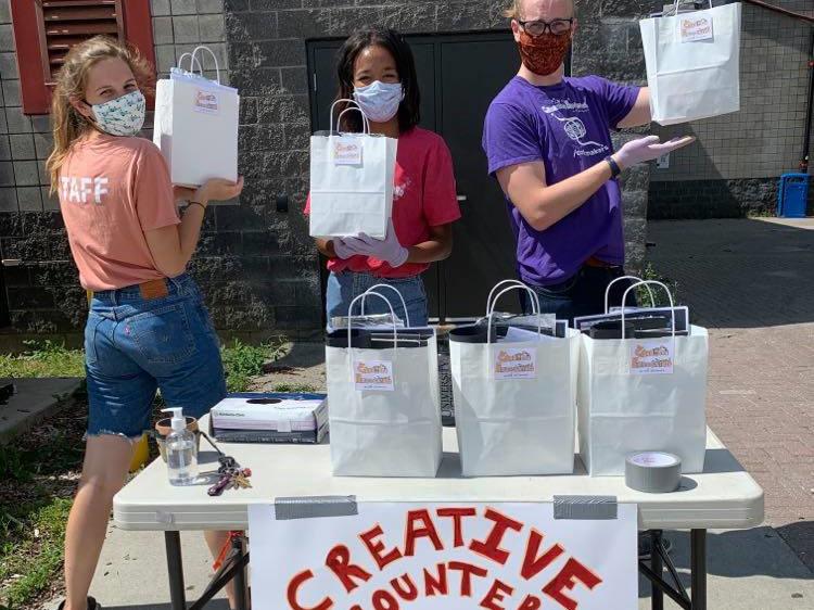 Staff standing behind Creative Encounters pickup table holding camp kit bags