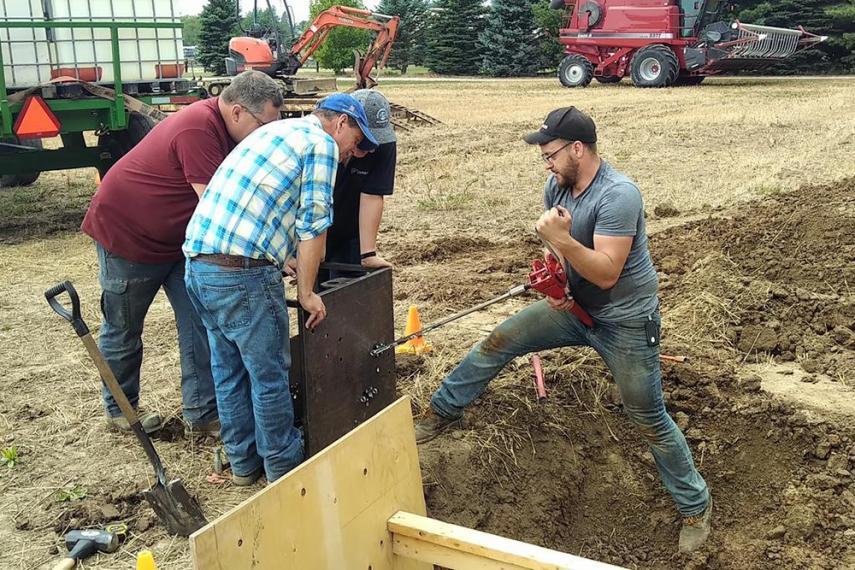 Alex Barrie giving soil demonstration to small group of farmers who are leaning in towards demo. Farming equipment in background.