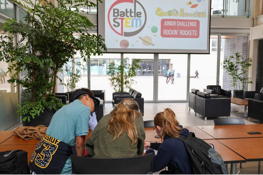 Three students writing on paper with a "BattleSTEM Junior Challenge Rockin' Rockets" sign projected above.