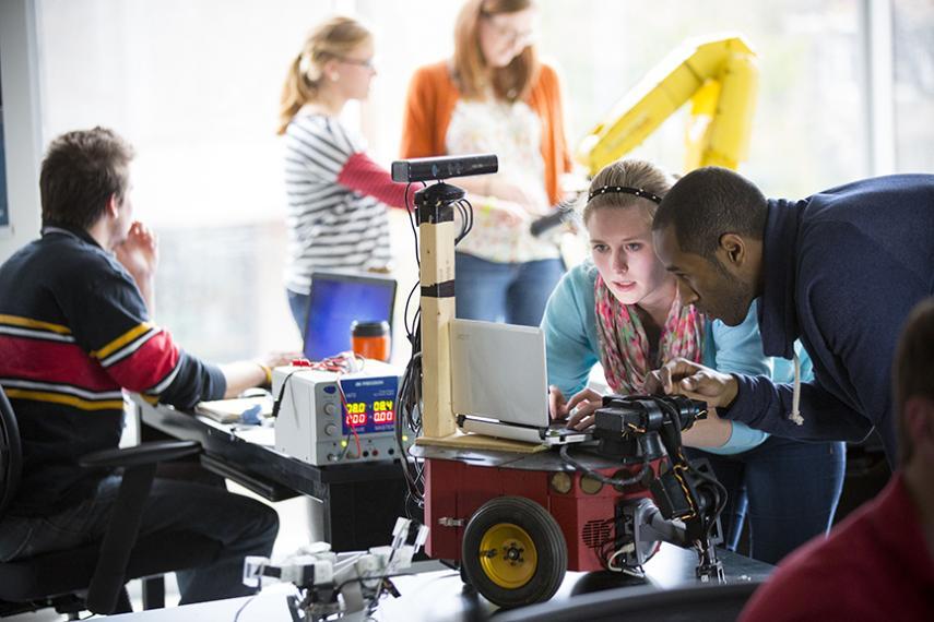 Two students assess technology in the foreground. In the background, other students are collaborating and working together