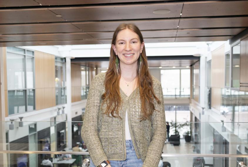 Portrait image of Mya SImpson standing on balcony above Thornborough atrium. 