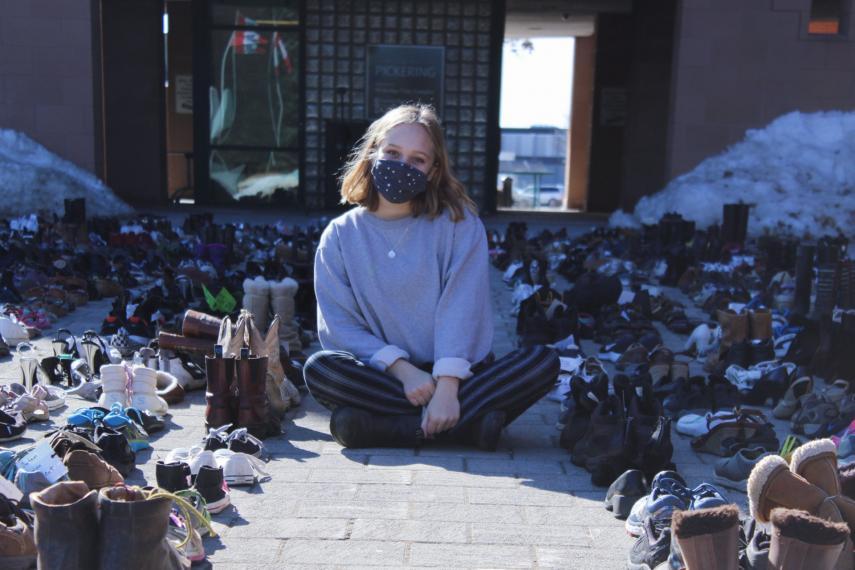 Ally Zaheer sits cross-legged wearing a mask and surrounded by hundreds of pairs of shoes outside city hall in Pickering