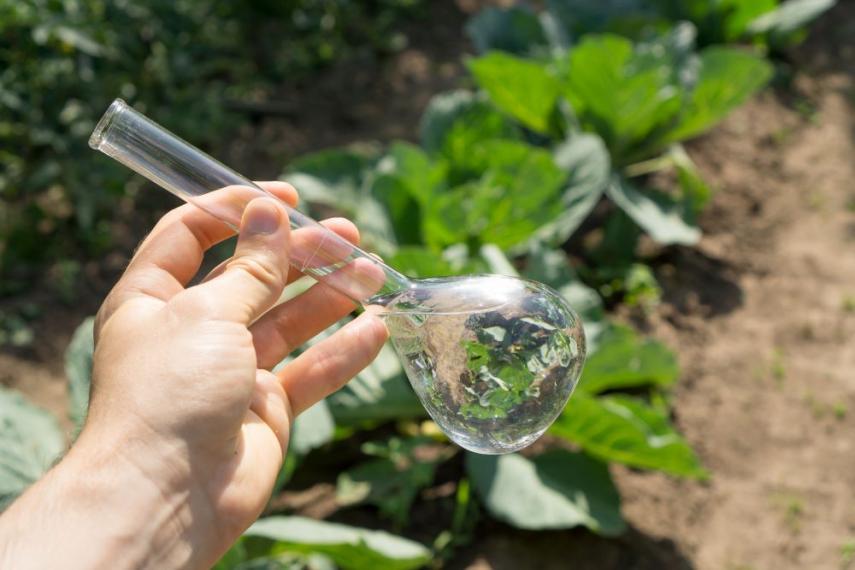 A hand holds a test tube with water. In the background is a farmer's field.