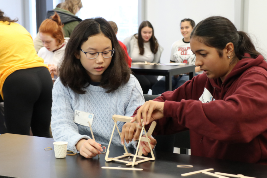 Two young girls making a device out of popsicle sticks with some people blurred in the background