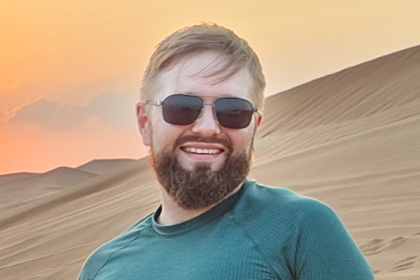 Man smiling at camera with sunglasses on and desert sunset in background.