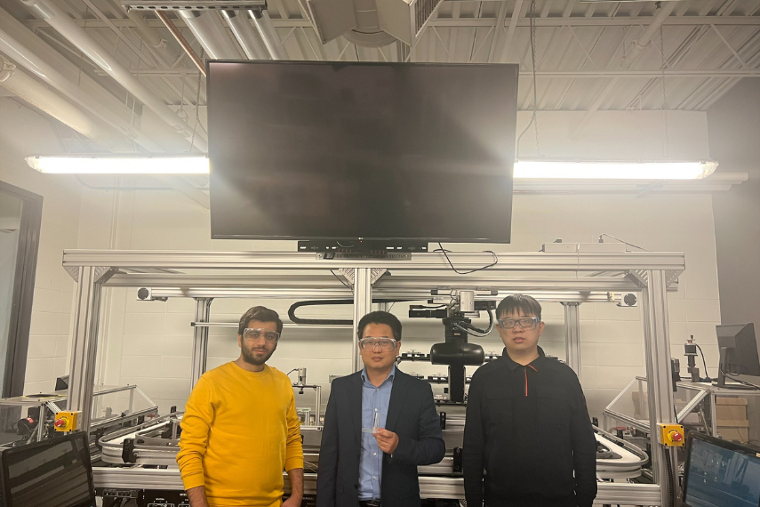 three men stand infront of large TV and technology with safety goggles on. 