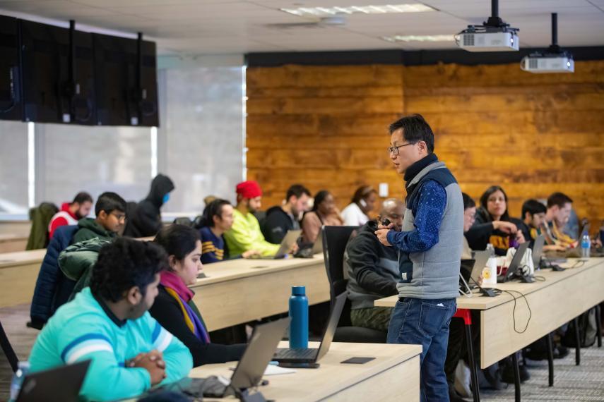 A professor speaks to a large classroom of students all paying attention with laptops in front of them.