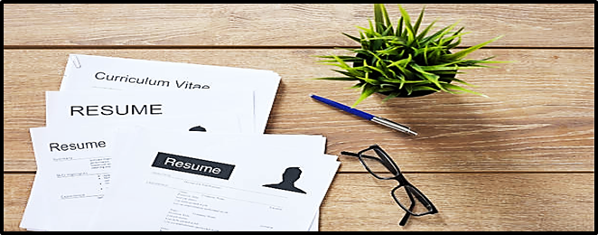 Pieces of paper lay on a wooden desk next to a plant, a pair of glasses, and a pen