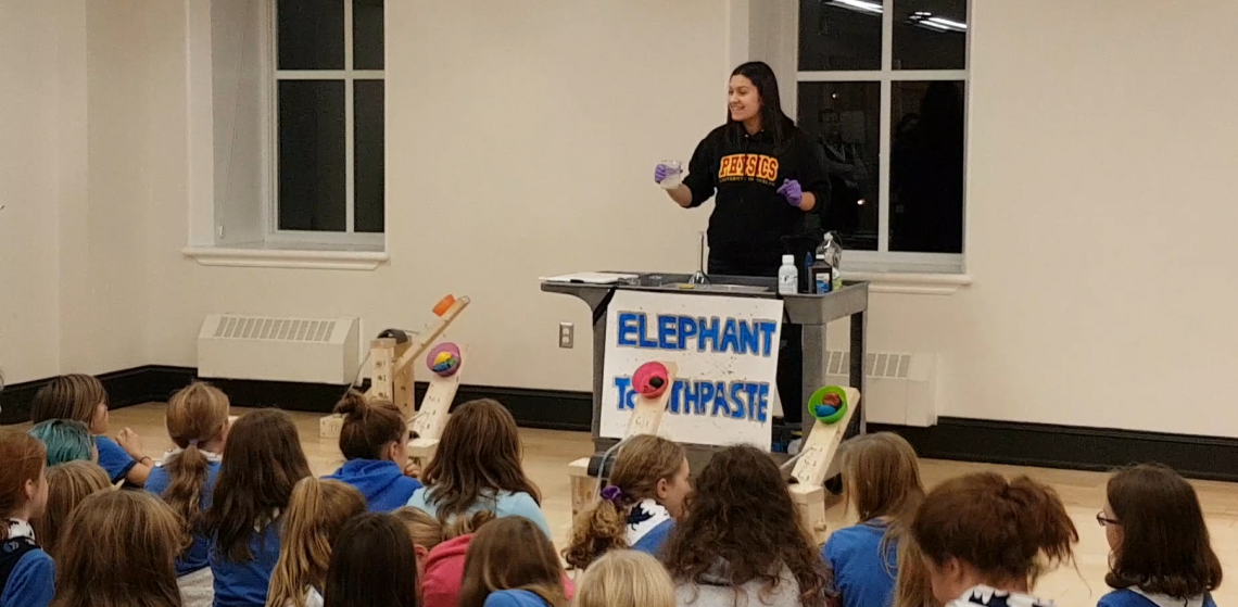 A photo of a woman standing at a podium conducting a science experiment in front of a room full of children. 