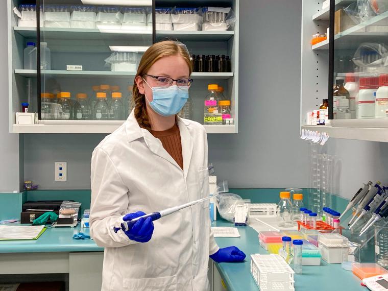 Jasmine Sollen stands in a lab surrounded by lab equipment while wearing a lab coat and full personal protective equipment. She holds lab equipment.