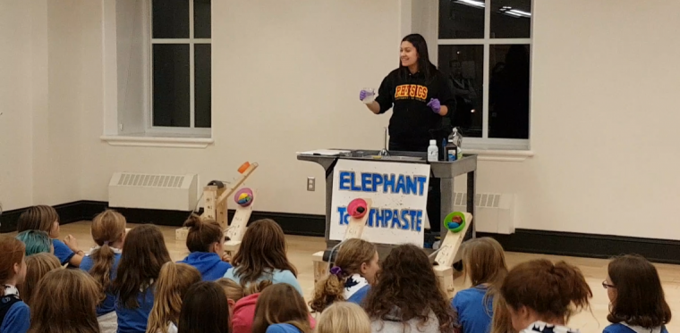 A photo of a woman standing at a podium conducting a science experiment in front of a room full of children. 
