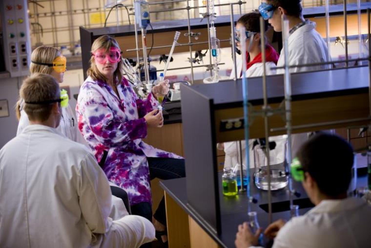 Bonnie Lasby sits in a lab and shows students a science demonstration. They are standing around her in personal protective equipment