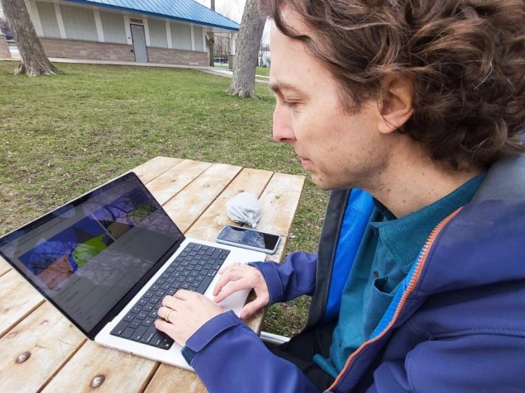 Graham Taylor sits at a wood park bench an works at a laptop