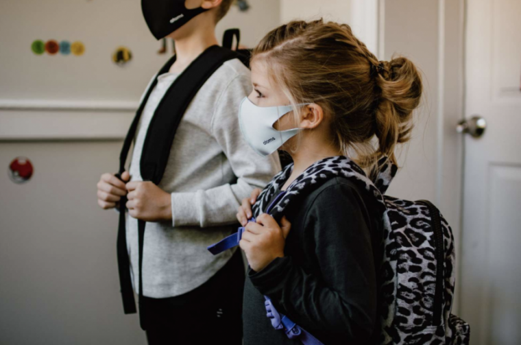 School-age student wears a mask and has a backpack on whilst standing in a classroom