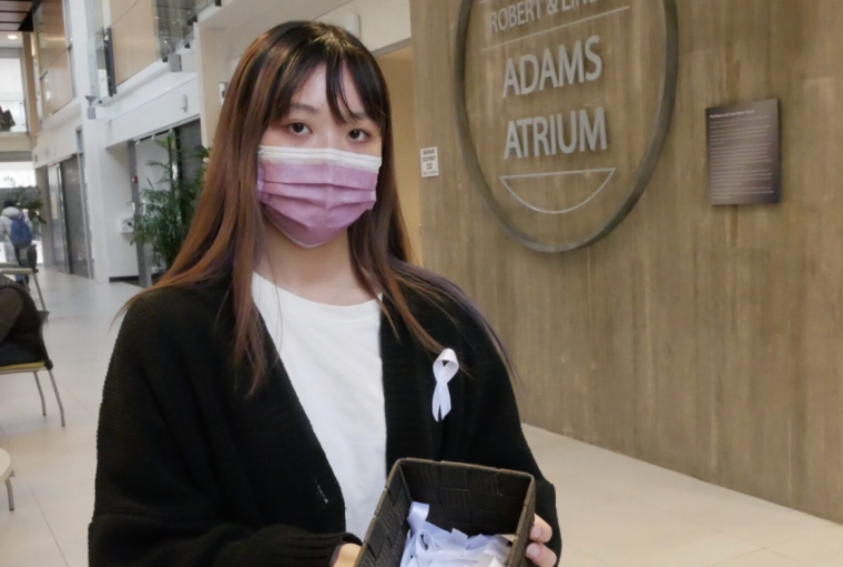 Grace Ly wears a mask in the Thornborough Building Adams Atrium while holding container of white ribbons for Dec 6 event