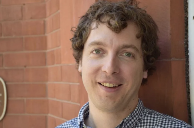 A picture of a man smiling in front of a brick wall 