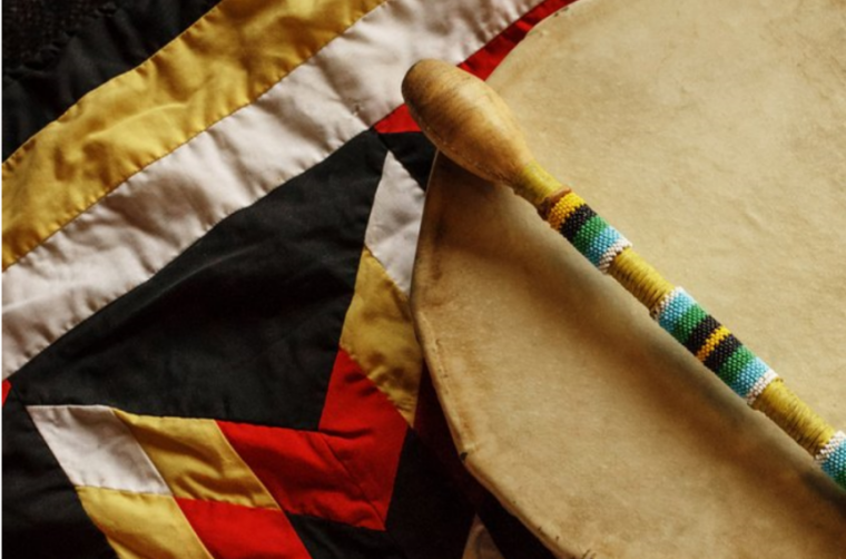 A rawhide hand drum and beaded drumstick sit on a star blanket made of red, yellow, white and black fabric.