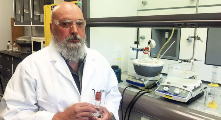 Man in a lab wearing a lab coat and goggles looing at the camera 