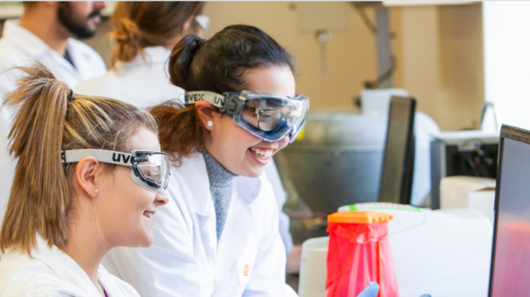 In a classroom laboratory, two students in lab coats, goggles and gloves watch a computer screen, smiling together.
