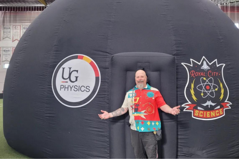 Orbax in bright colourful shirt standing in front of portable planetarium. 