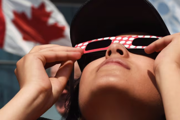 Kid looking up at solar eclipse with glasses on and Canada flag in background. 