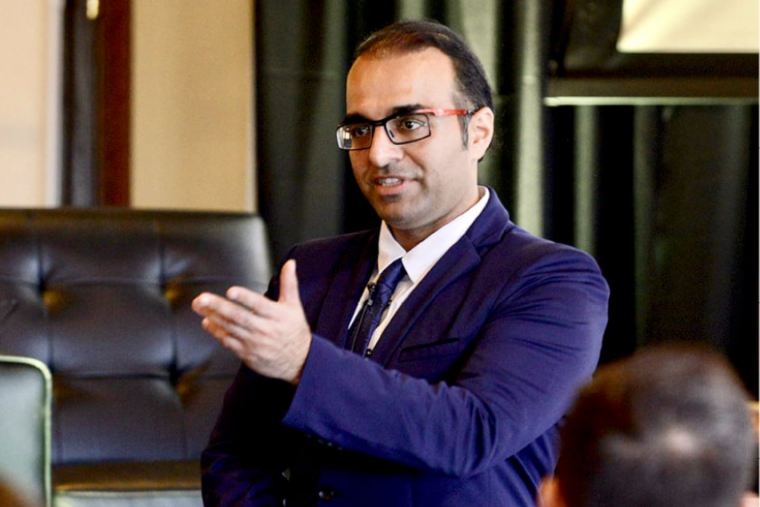 Dr. Ali Dehghantanha speaking to a crowd with his arm raised, wearing a dark blue suit.