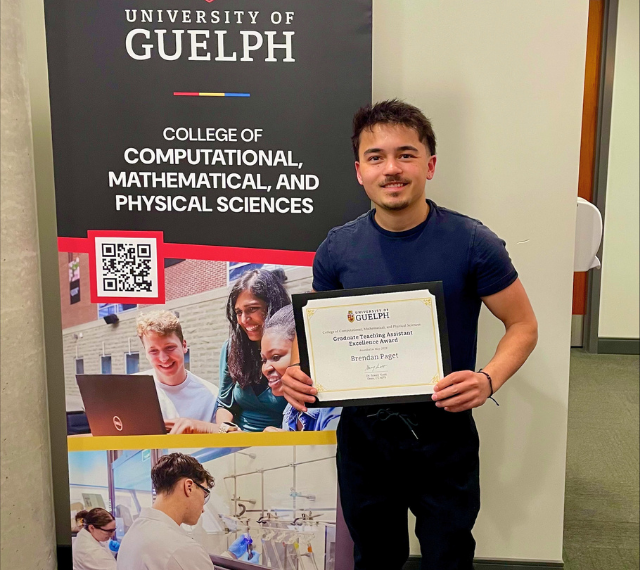 Brendan Paget stands infront of a Guelph banner holding his Teaching Assistant Excellence Award.