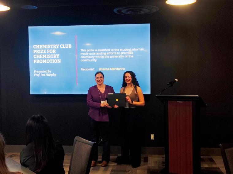 Professor Jennifer Murphy stands with Brianna Mandarino holding a black folder infront of a blue screen with the award detail listed.