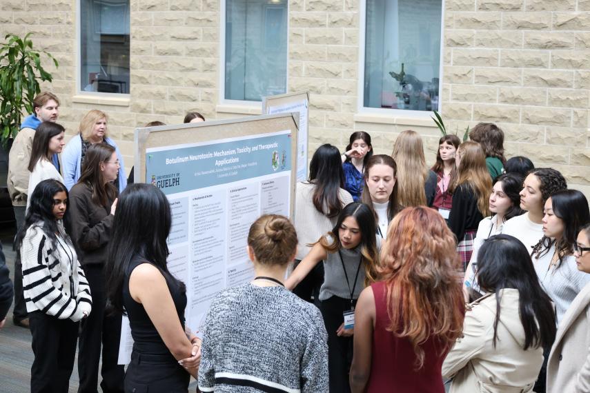 A group of symposium attendees gather around a scientific poster as the presenter explains the poster.
