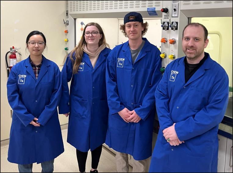 Four TAs stand in front of fumehoods in a laboratory. Each TA is wearing a blue lab coat.